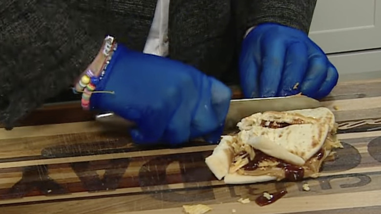 Gloved hands slicing a sample of Jeff Daniels' favorite sandwich on top of wooden chopping board.