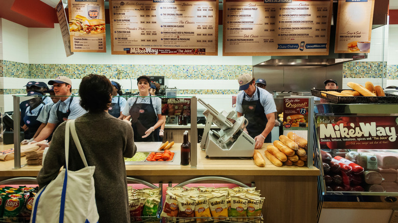 Customer at Jersey Mike's counter with workers behind it and bread stacked near the slicer