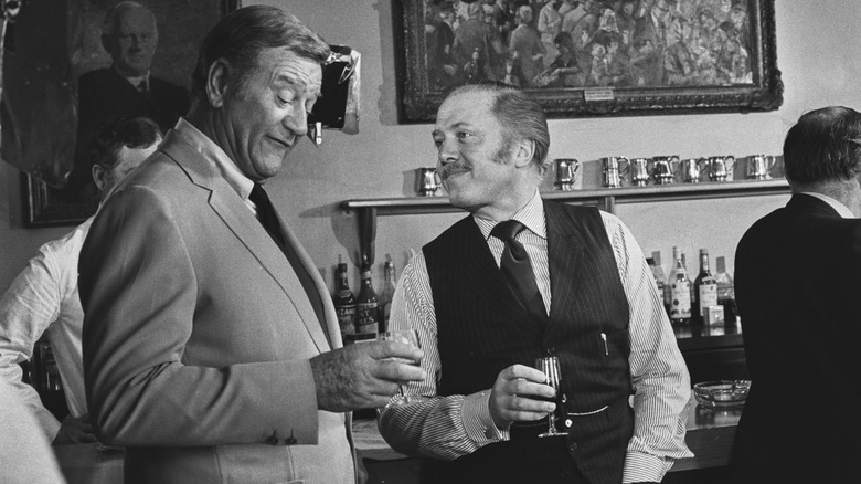 John Wayne in a black and white still from a movie set, drinking at a bar