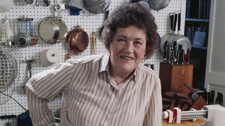 Julia Child sitting in a kitchen with her various tools, pots, and pans behind her.