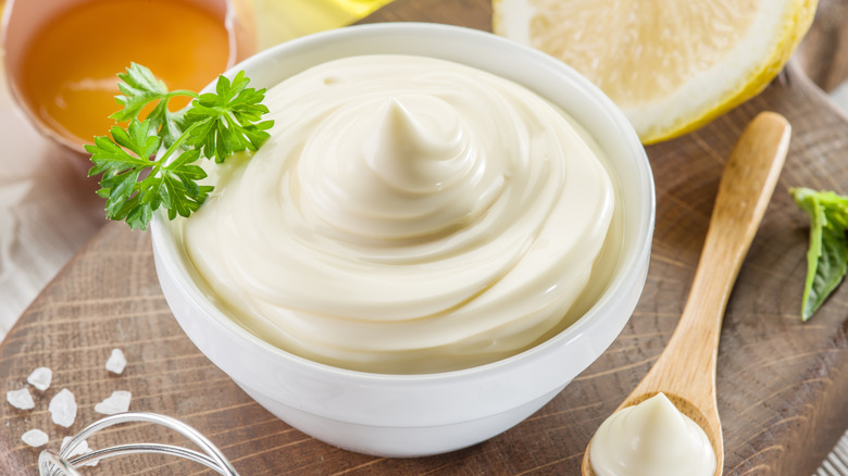 A bowl of mayonnaise on a wooden surface neear a spoon and mayo ingredients