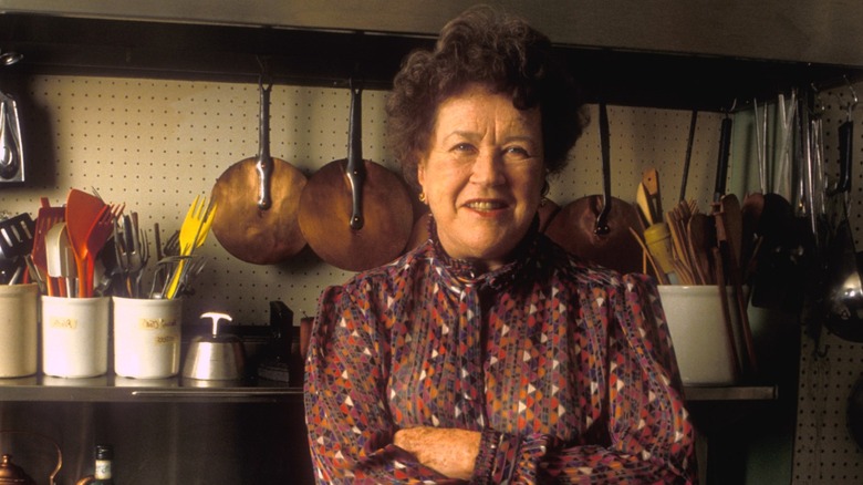Julia Child standing before a shelf full of cookware.