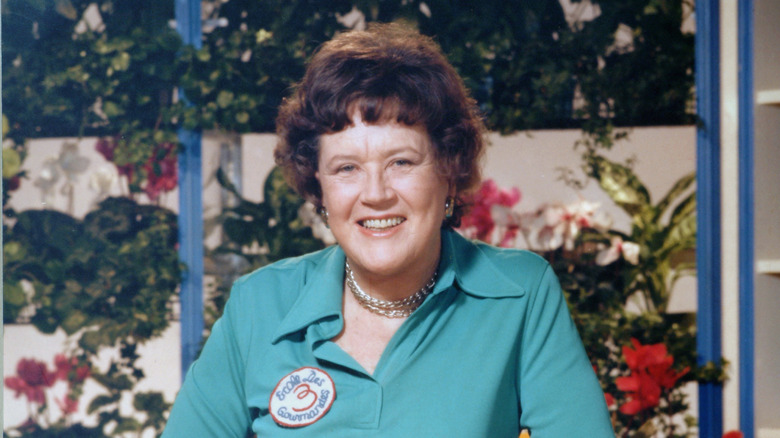 Julia Child standing in her kitchen wearing a badge from her cooking school.
