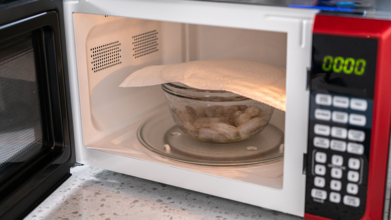 A glass bowl of shrimp covered with a paper towel sitting in the microwave.