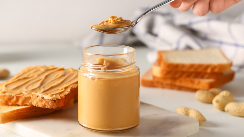 Someone taking a spoonful out of a jar of peanut butter with sandwich bread in background