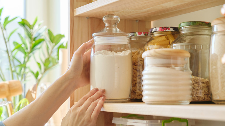 A person placing a glass jar of flour on a pantry shelf.