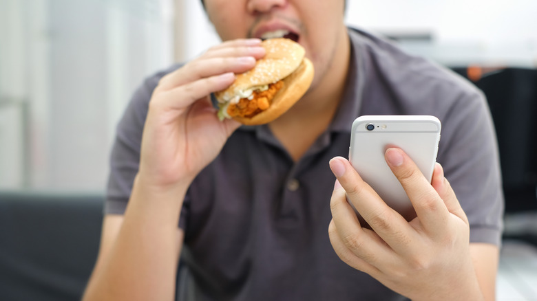 A person checking a phone while eating a chicken sandwich.