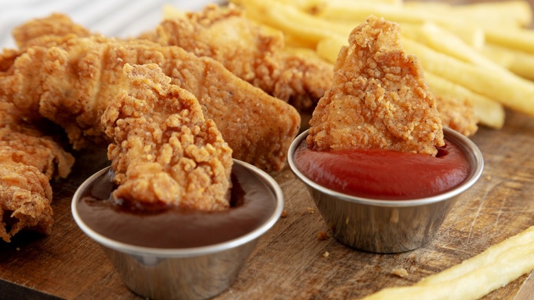 Chicken tenders dipped into sauce cups on wood board with fries.
