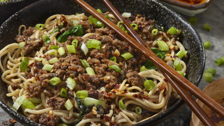 A bowl of dan dan noodles with meat, green onions, and chopsticks