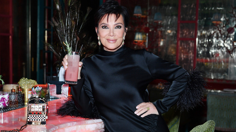 Kris Jenner wears a black dress and holds a pink cocktail by a pink marble bar