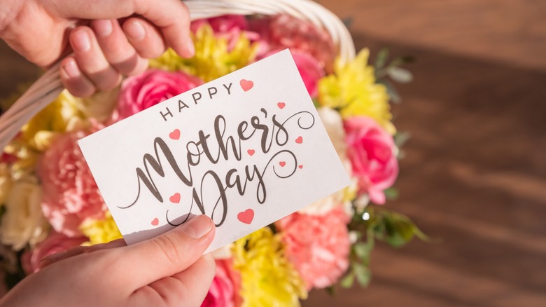 Hand holds a card reading Happy Mothers Day and holds a gift basket with blooming carnations