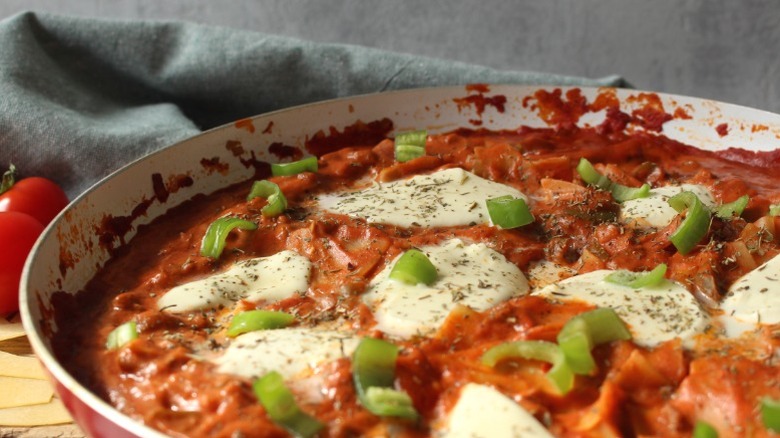 A skillet of one-pot lasagna sitting on a counter with tomatoes and a dish rag nearby.