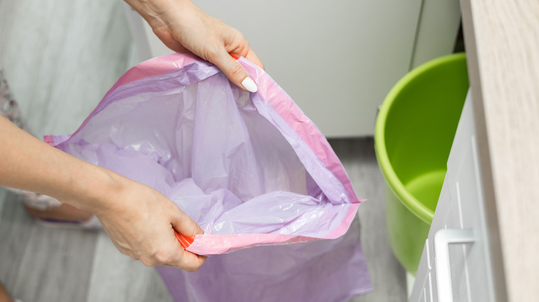 A person with holding a trash bag open ready to put it inside a trash can