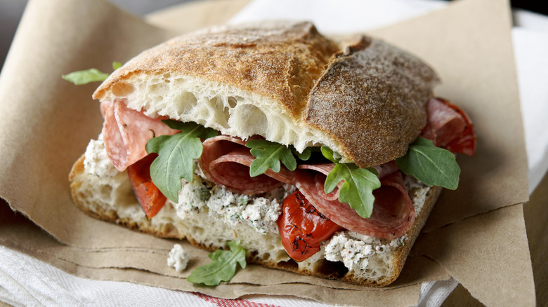 A crusty sandwich roll with deli meats, tomatoes, and greens on sandwich paper.