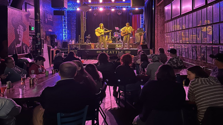 Two musicians playing on stage in a narrow room while patrons sit at wooden tables with beers on them
