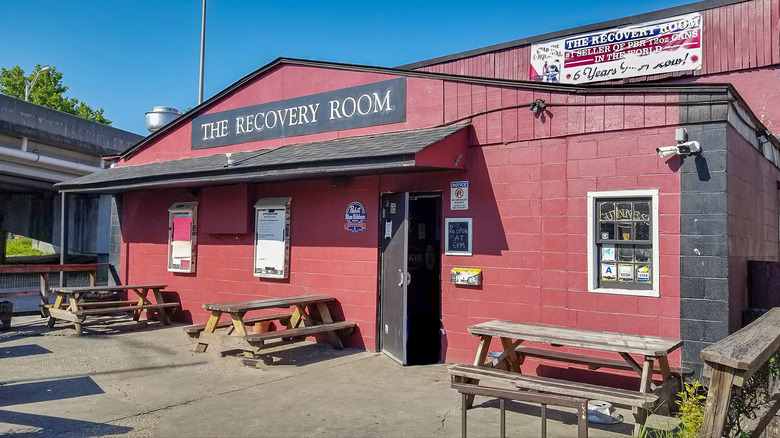 Red cinder block building with picnic tables outside and a plain black and white sign that says The Recovery Room