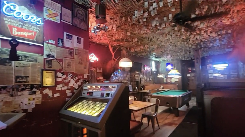 A jukebox and a pool table in a dive bar with a ceiling plastered with dollar bills