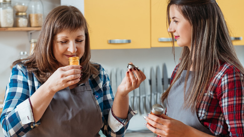 Two women smelling kitchen spices in the kitchen with aprons on.