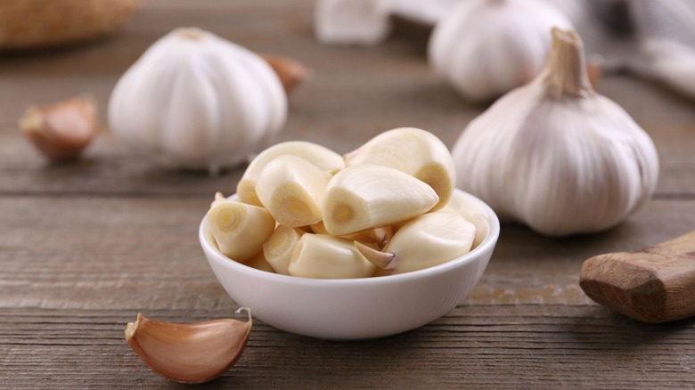 Peeled garlic cloves in white bowl with whole bulbs in background on wood table