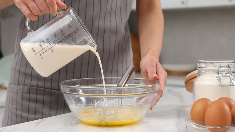 Woman pouring milk from a measuring cup into bowl of raw eggs with whisk on a white marble table, closeup