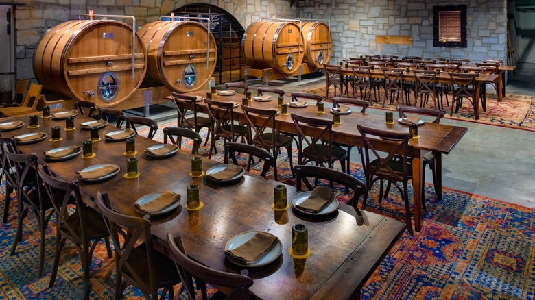 A large room with long wooden tables and eclectic carpets as well as brewing casks against one wall