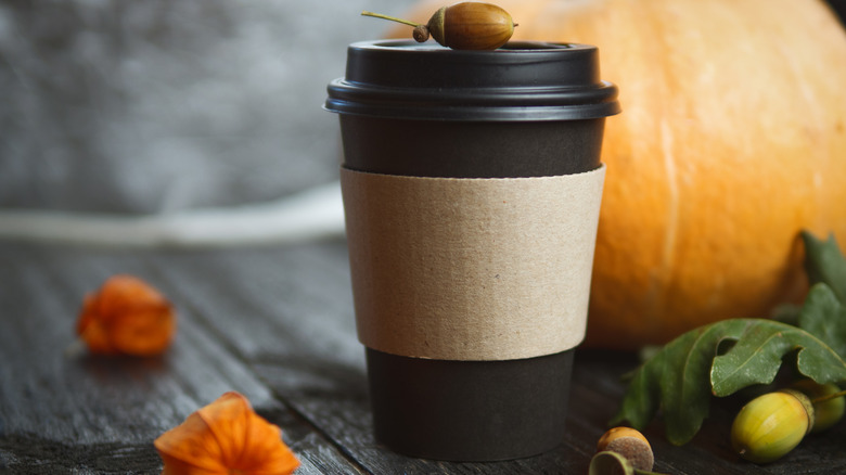 A black plastic to-go coffee cup on a wooden surface with acorns and a pumpkin