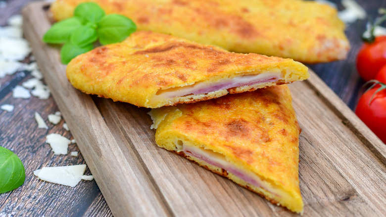 Close up of a homemade hot pocket sliced in half with ham and cheese filling on a wooden cutting board, with various ingredients surrounding.