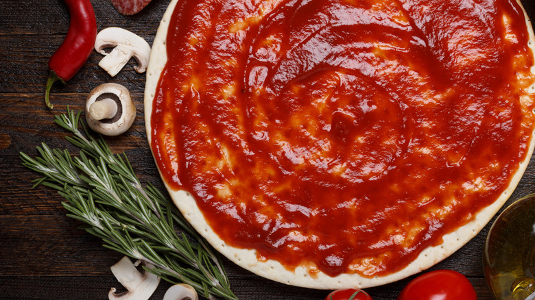 Fresh pizza dough spread with pizza sauce alongside rosemary, mushrooms, olive oil, and chile pepper