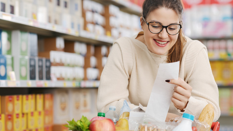 A young woman looking at her grocery receipt leaning over her cart full of groceries