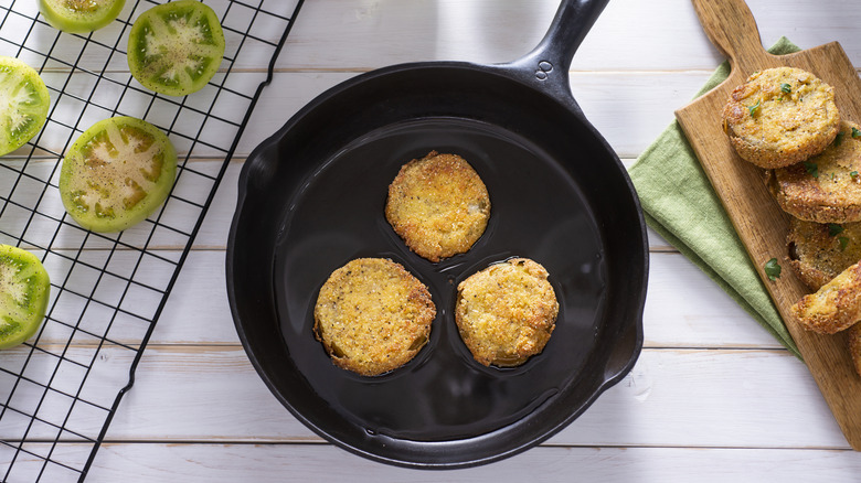 Fried green tomatoes in a pan with unbaked tomatoes on a grate along side