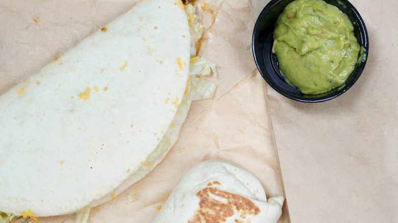 A taco bell taco and burrito on paper next to a side of guacamole sauce