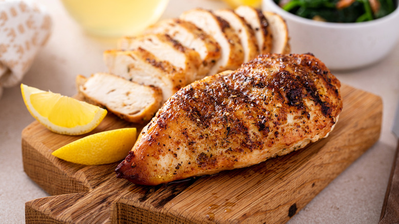 Grilled chicken with lemon slices beside it on a thick wooden cutting board
