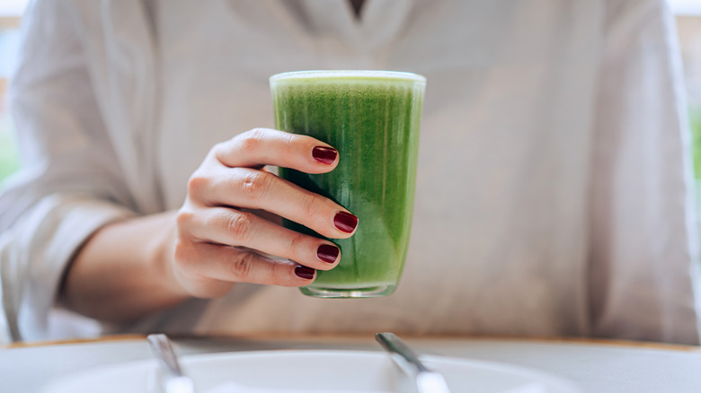 Close-up, mid-section of woman holding a glass of fresh and healthy green juice. She has dark red nails and is wearing a light colored long sleeve shirt.