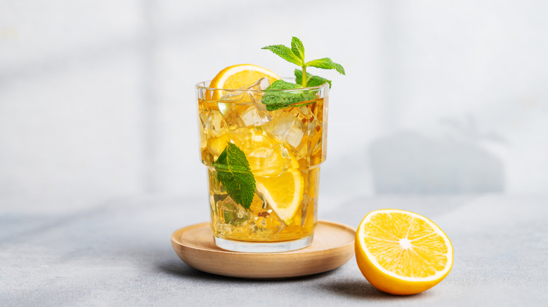Iced tea with lemon and mint in a glass atop a wooden coaster and a sliced lemon on the side.