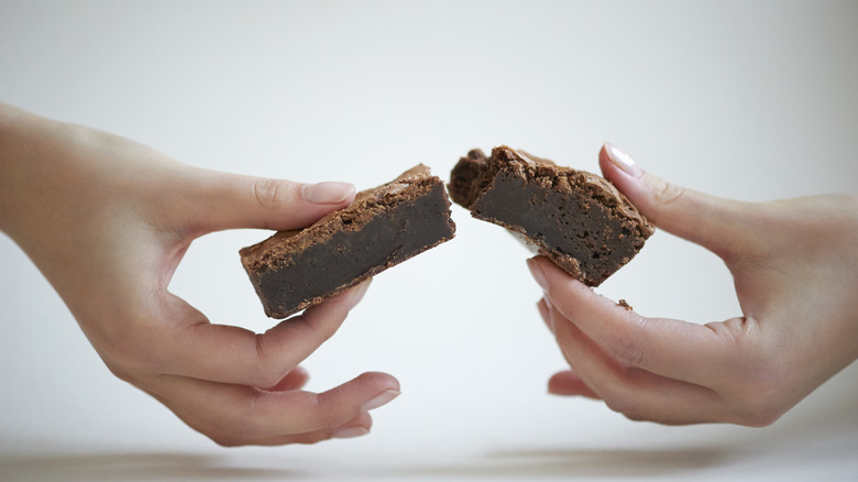 Close up of female hands breaking a home made chocolate brownie in half against a white background.