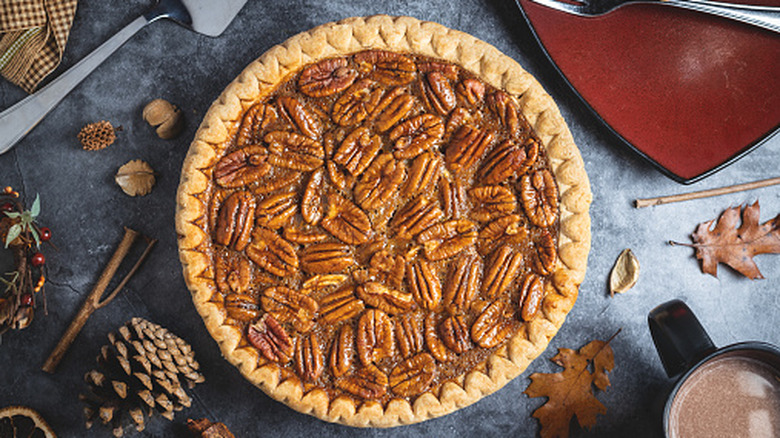 Top down view of a fresh whole pecan pie on a table with rustic fall leaves, an empty plate, and cup of hot cocoa nearby.
