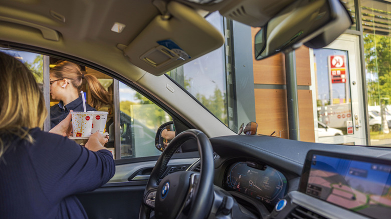 A person in a car receives drinks at a McDonald's drive-thru window from an employee, with the interior of the car and the restaurant in view