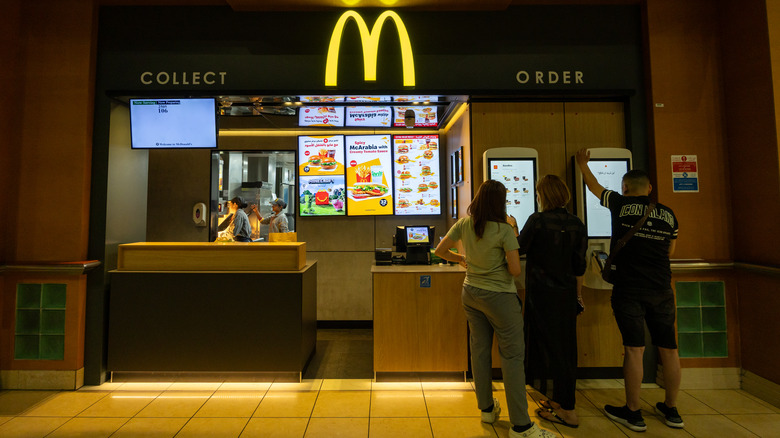An interior view of a McDonald's ordering area, with people at digital kiosks and workers in the back