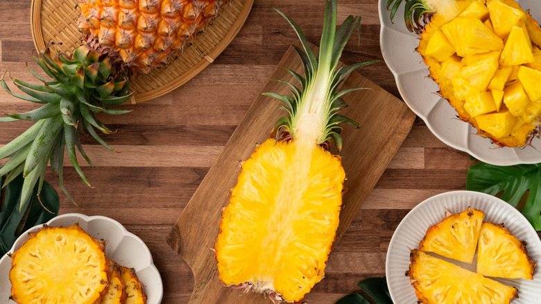 Juicy pineapple chunks and slices arranged around a halved pineapple with a wooden cutting board and tropical leaves in the background.