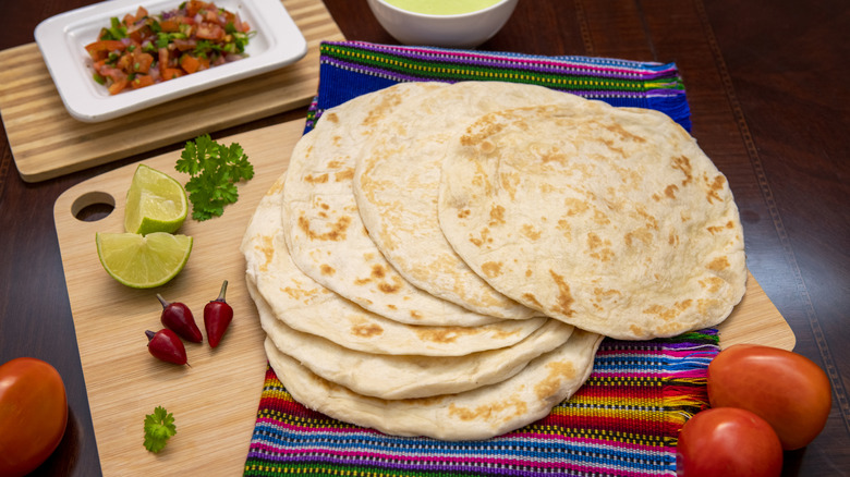 Large tortillas spread out in a fan on a colorful cloth, surrounded by tomatoes, lime pieces, peppers, and herbs