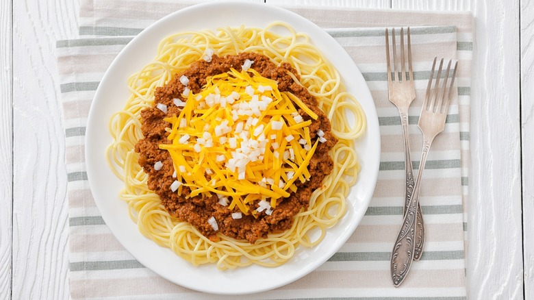 Cincinnati chili, spiced meat sauce, chopped onion, grated cheddar cheese on top of spaghetti on plate on white wood table with forks, american cuisine, flat lay, close-up, not AI generated