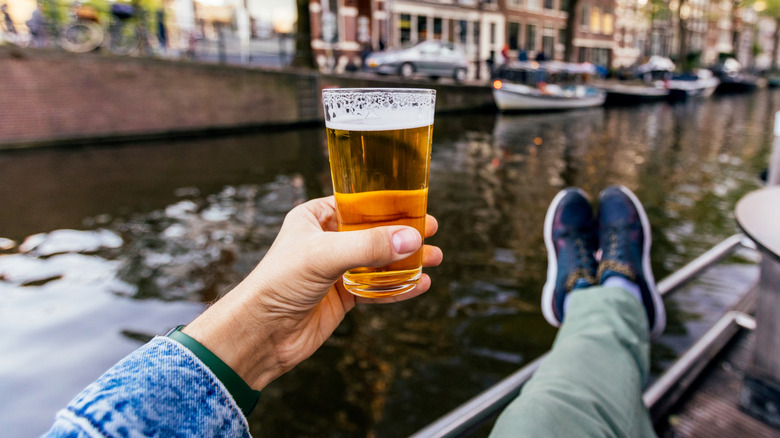A person holding a glass of beer in their hand in front of a river.
