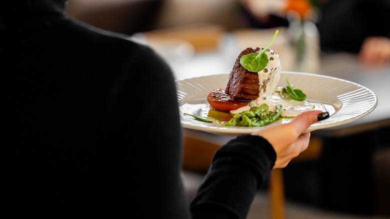 An elegantly plated gourmet dish, possibly a beef fillet with roasted vegetables and a creamy sauce, is being held by a server in a fine-dining restaurant