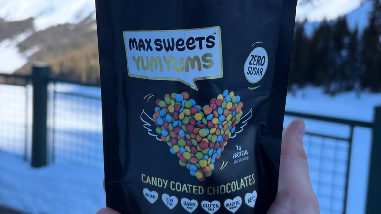Package of Max Sweets Dark Chocolate Sugar Free YumYums being held by a hand with snow-covered mountains in the background