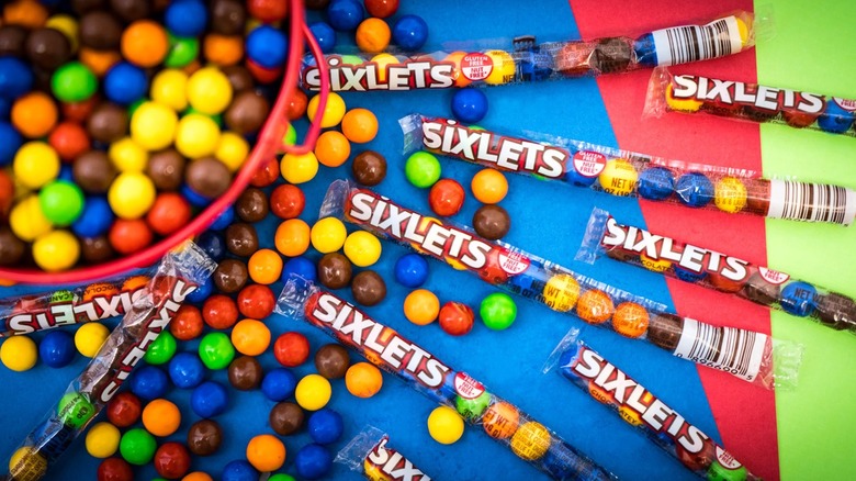 Numerous Sixlets packets surrounded by scattered, loose Sixlets and a bucket of loose Sixlets