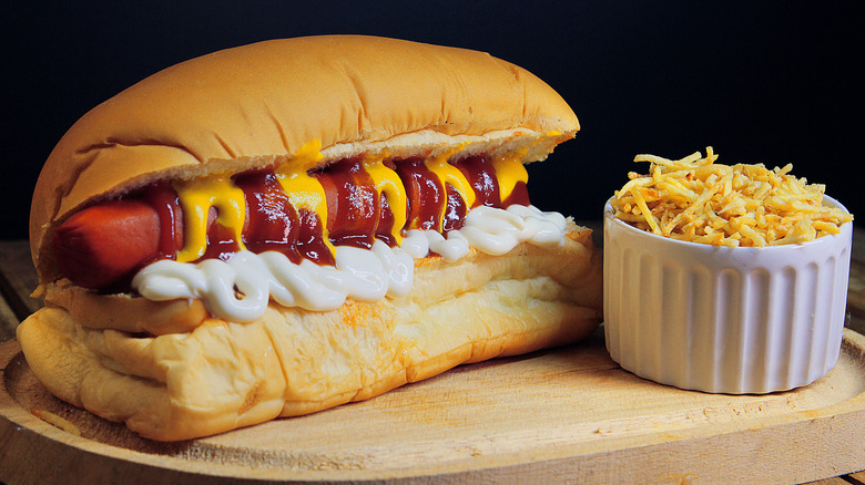 Hot dog in a bun on a wooden cutting board with mustard, ketchup, and mayo next to a cup of fried onions