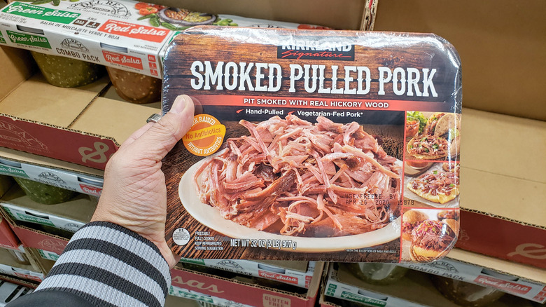 A hand holds a package of Kirkland Signature smoked pulled pork to the camera in a Costco warehouse.