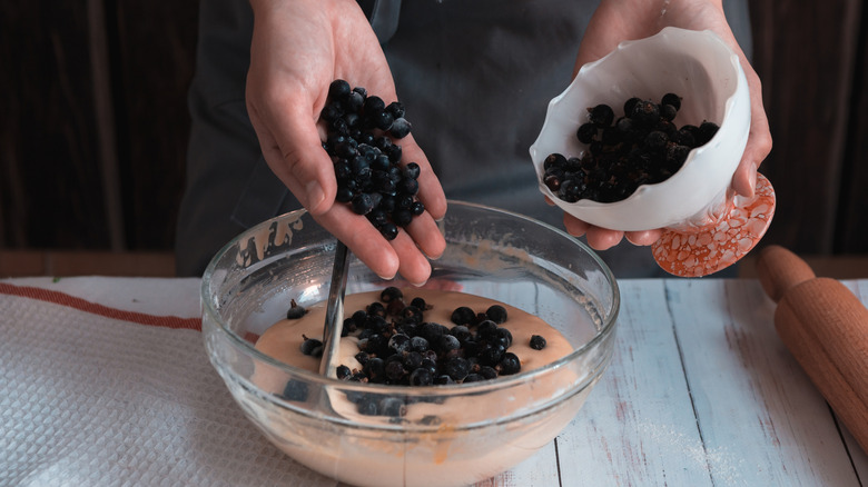 A person adds fresh blueberries to muffin batter from a dish