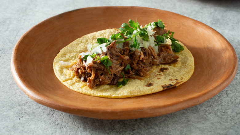 Tortilla filled with shredded meat and topped with cilantro and white onion on an earthenware plate.