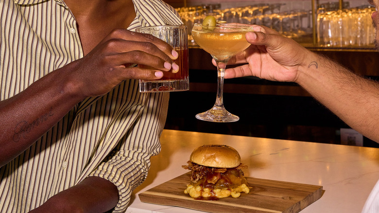 Two people raising cocktails over a burger at R House Wynwood bar